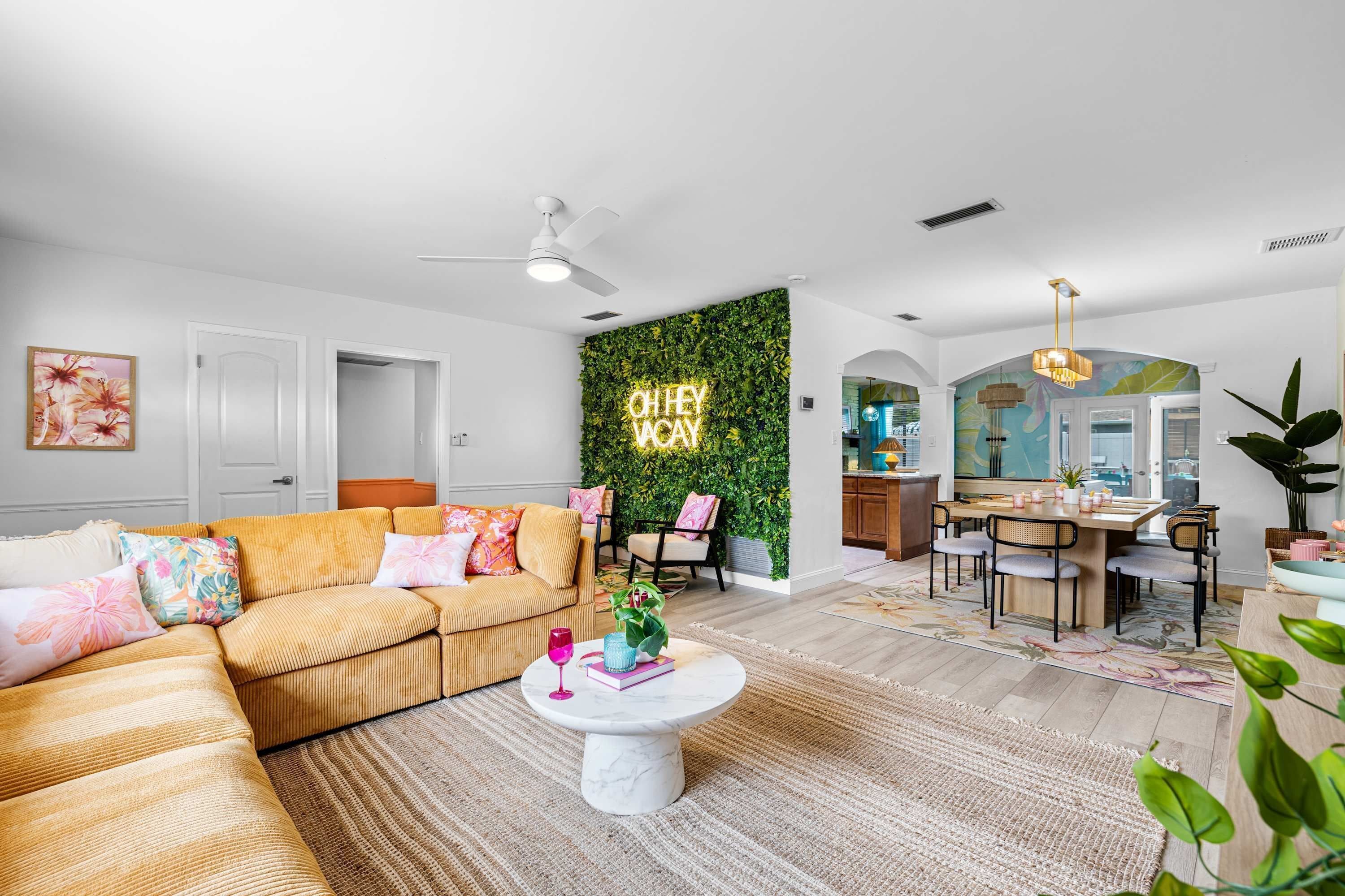 Una sala moderna con un sofá, una mesa de centro y una pared decorada con un diseño de hojas verdes. Hay un comedor con sillas y una mesa.