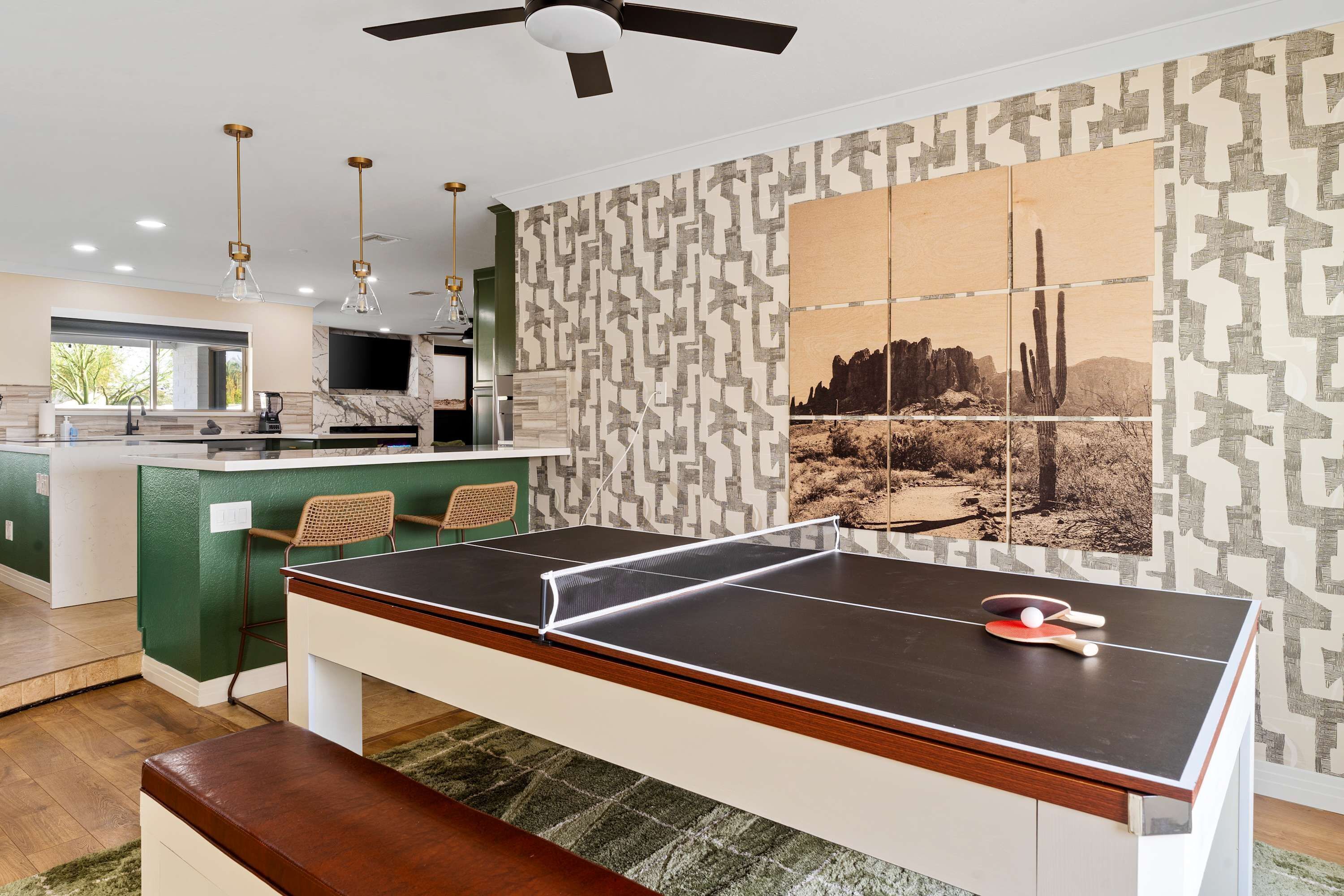 Uma mesa de pingue-pongue com um banco, uma parede verde com design estampado e uma área de cozinha com televisão e iluminação moderna.
