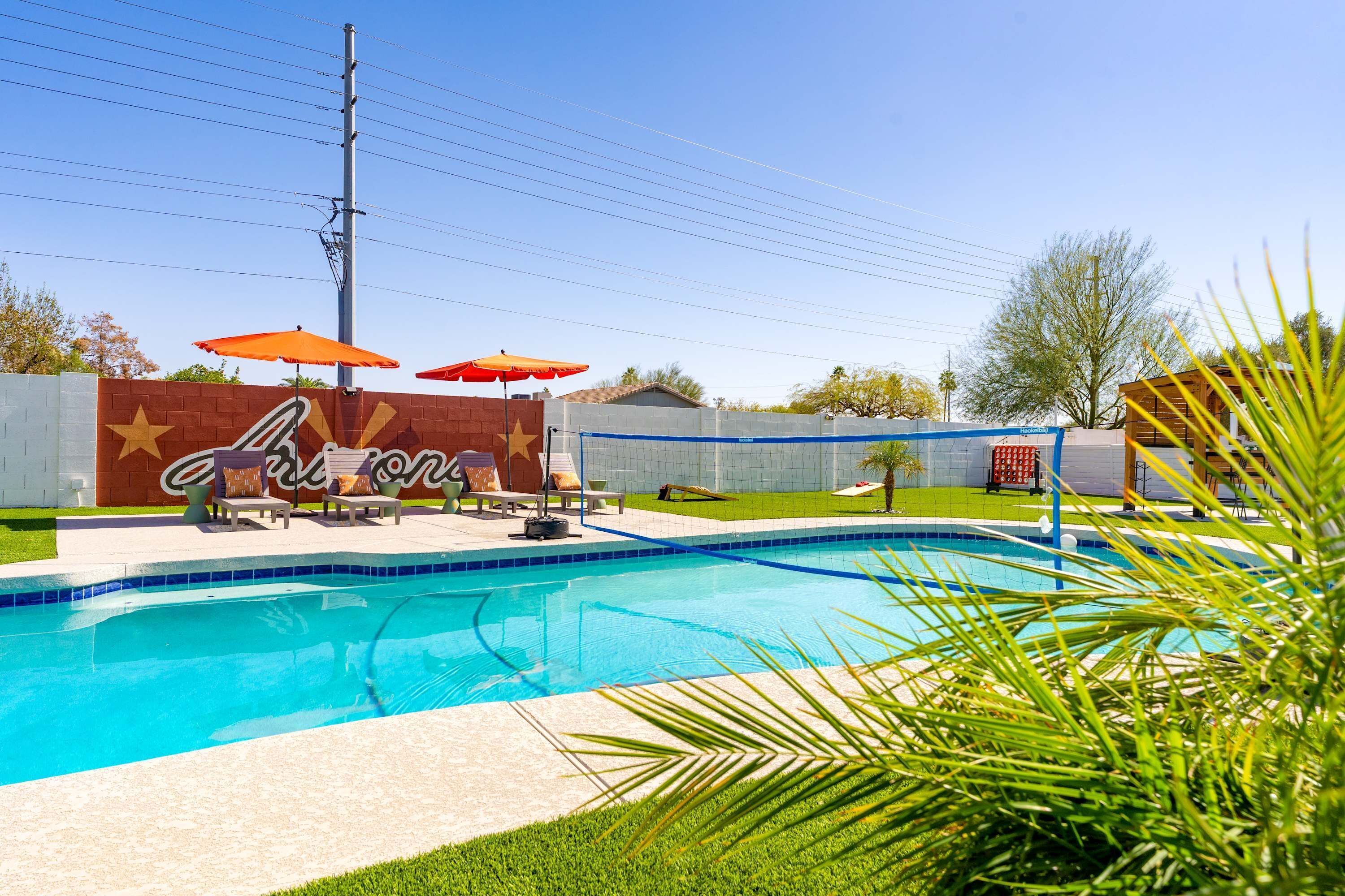 A swimming pool with lounge chairs and umbrellas, surrounded by a brick wall with a mural, and a clear sky.