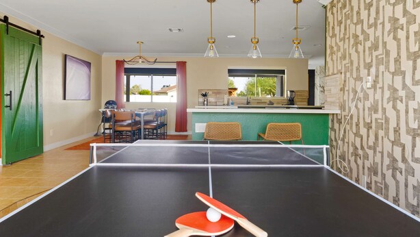 A ping pong table with a paddle and ball, a dining area with chairs, a kitchen counter, and a hanging light fixture. - The Ridge Family Pickleball Pool Hot Tub (Phoenix)