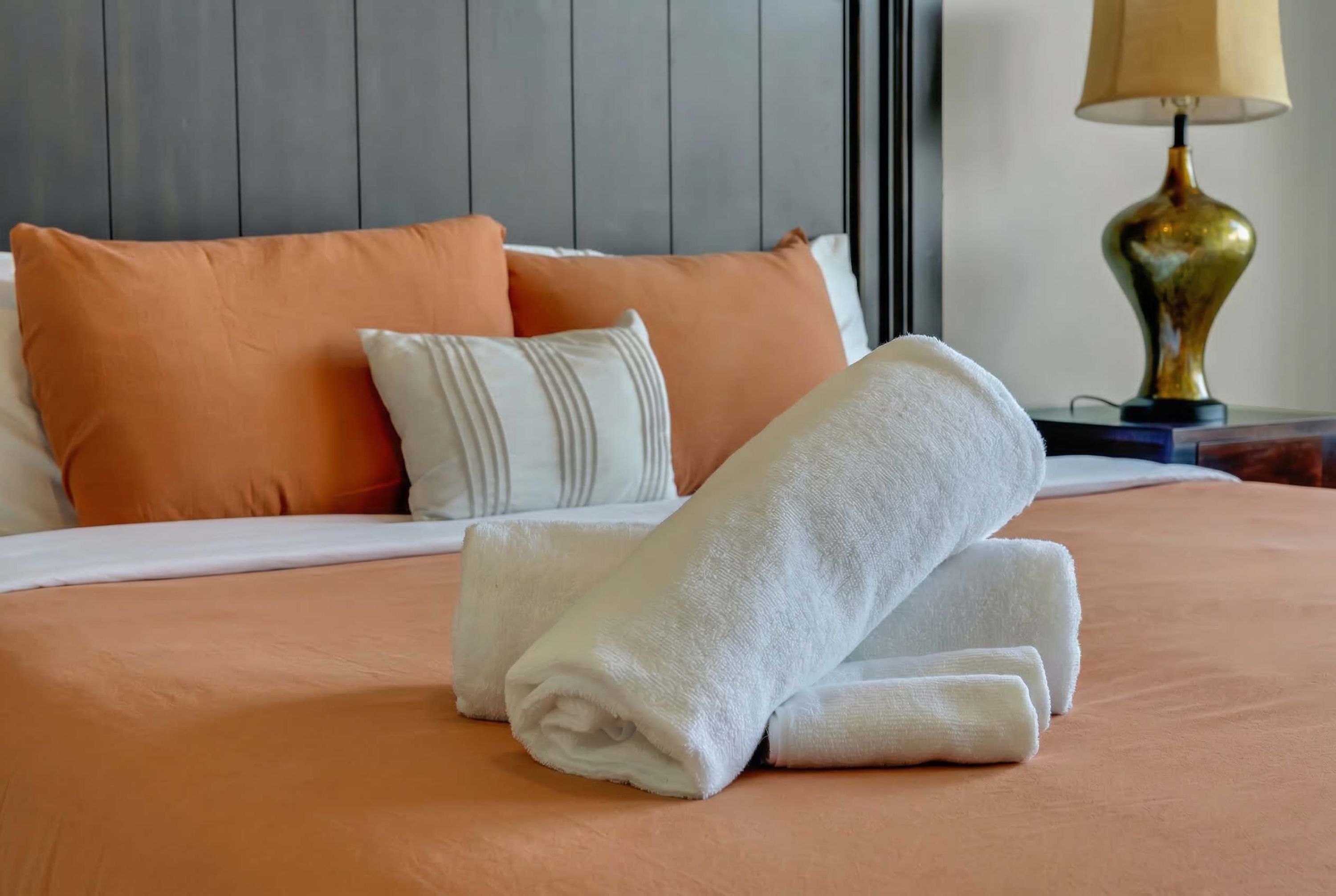 A neatly made bed with a folded white towel and two pillows, one orange and one white, in a room with a grey headboard and a lamp on a bedside table.