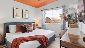 Een slaapkamer met een bed, kussens, een nachtkastje, een lamp, een raam met gordijnen, een hanglamp en een vaas met gedroogde bloemen.