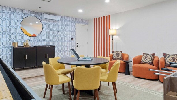 Een moderne woonkamer met een ronde tafel, gele stoelen, een oranje bank en een behang met patroon.