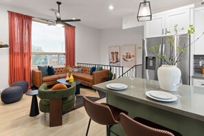 Uma sala de estar moderna com sof á, mesa de centro e pufes. Há uma área de cozinha com geladeira e uma mesa de jantar com pratos e um copo.