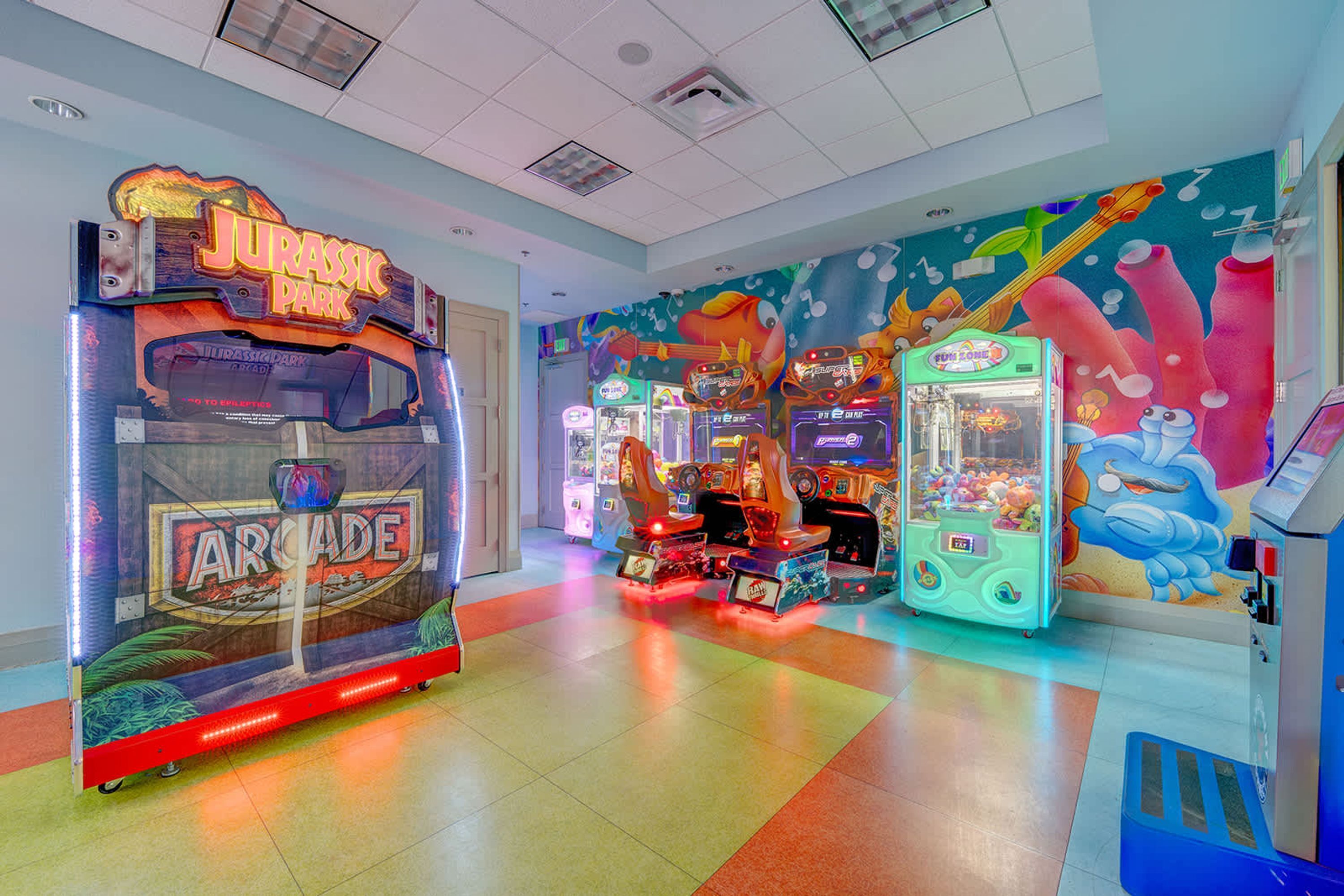 Une zone d’arcade bien éclairée, avec différents jeux, notamment un jeu sur le thème de « Jurassic Park » et un jeu de griffe sur le thème de « Raw Deal ».