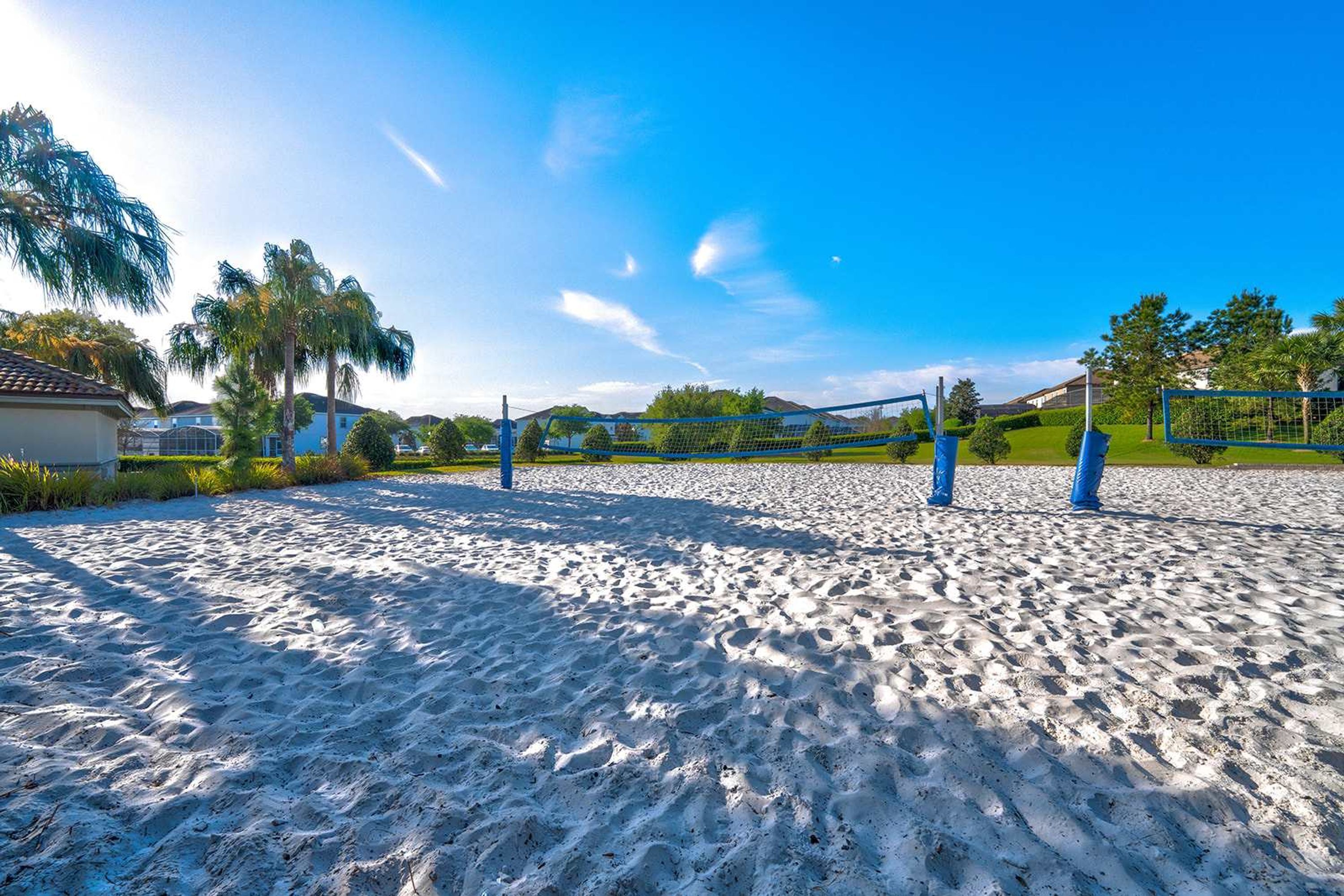 A sandy beach volleyball court with a net, surrounded by greenery and houses in the background.