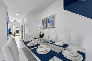 Un coin repas avec une longue table dressée pour quatre personnes, des chaises blanches et deux œuvres d’art encadrées accrochées au mur.