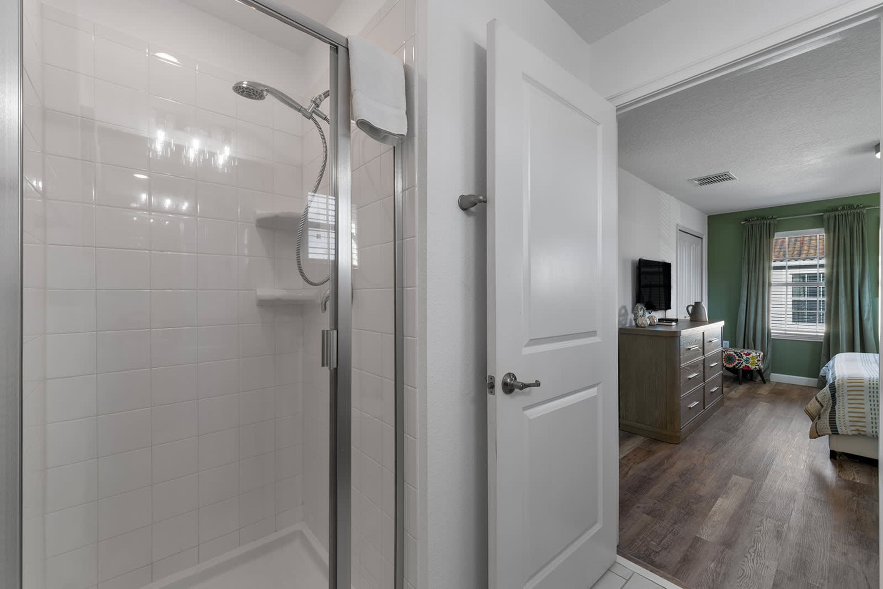 A bathroom with a glass shower enclosure, white tiled walls, and a white door leading to a bedroom with a wooden floor, a dresser, and a bed.