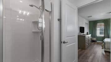 A bathroom with a glass shower enclosure, white tiled walls, and a white door leading to a bedroom with a wooden floor, a dresser, and a bed.