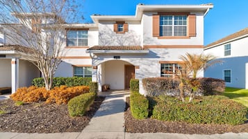 A two-story residential building with a central entrance, a covered patio area, and a well-maintained front yard with various shrubs and a tree.