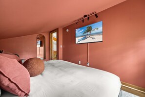A bedroom with a bed, a round rug, a painting of a beach scene, and a door leading to another room. - Birdie Creek Ranch - Private Golf Course Resort (Driftwood)