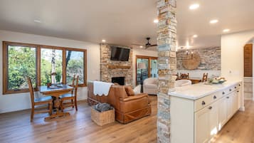A spacious living area with a fireplace, a stone accent wall, a large window with a view of greenery, and a kitchen with white cabinetry.