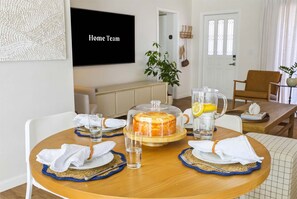 A dining area with a round table set for four, a cake on a stand, a pitcher with a lemon slice, and a television displaying 'Home Team' on the wall. - Minutes to AMI Pool Game Room Sleeps 11 (Bradenton)