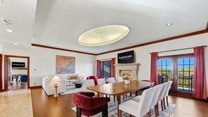 Un vaste espace dédié à la salle à manger, avec une longue table en bois, des chaises blanches et un fauteuil rouge qui apporte une touche de couleur. On y trouve une cheminée sur laquelle est accrochée une plaque, ainsi qu’une grande fenêtre ornée de rideaux.