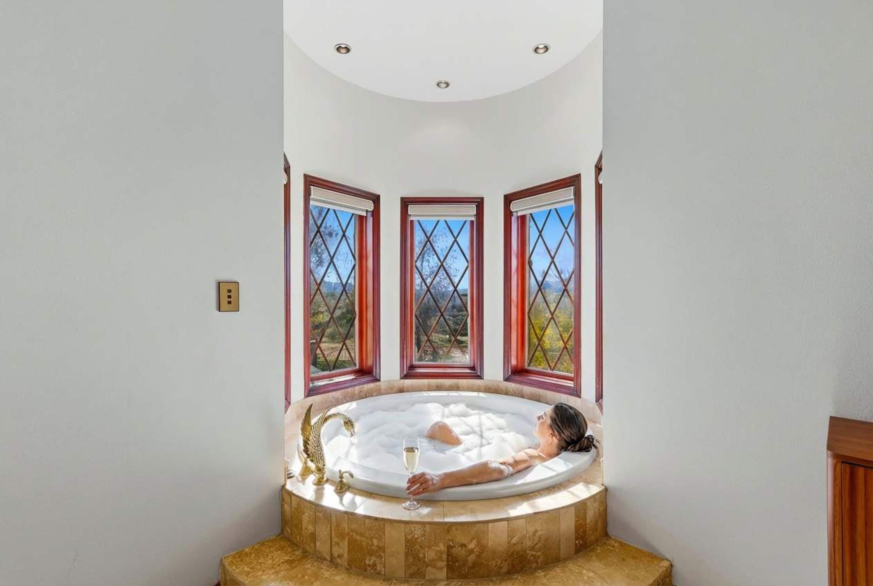 A person relaxing in a freestanding bathtub with a view of a mountainous landscape through three windows.