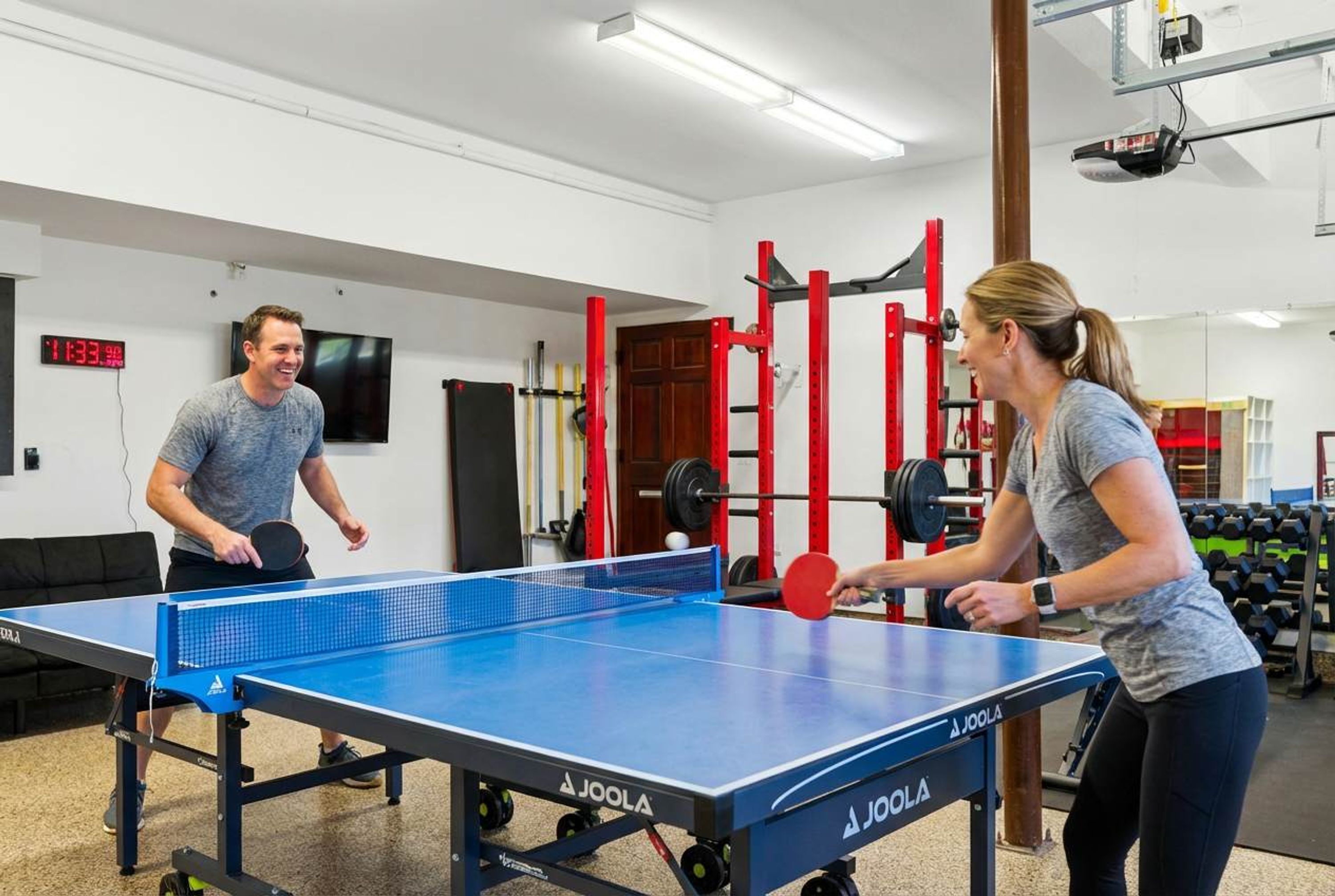 A ping pong table with two people playing, surrounded by gym equipment and a digital clock.