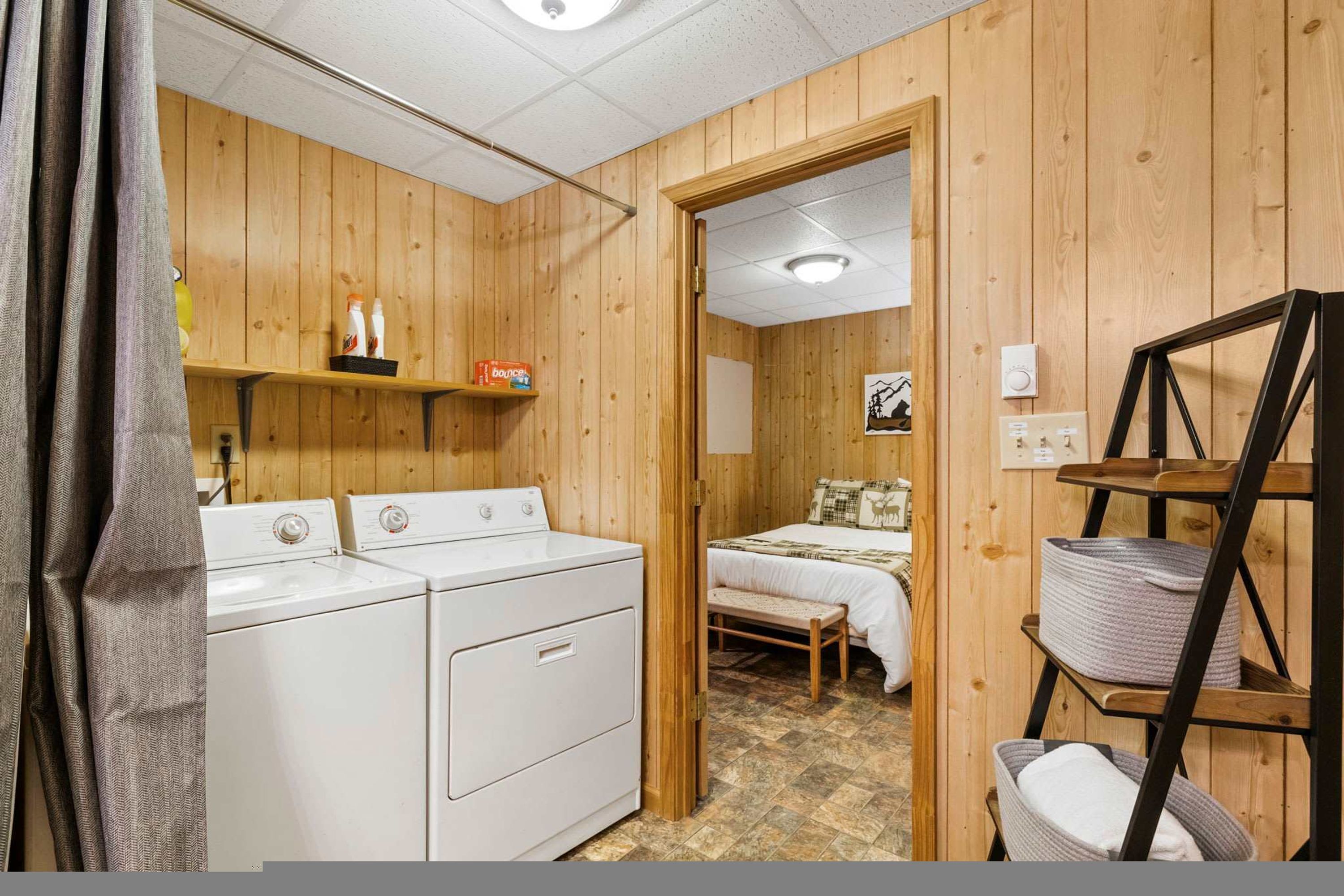 A room with a wooden interior, a washing machine, a bed with a checkered bedspread, and a shelf with toiletries.