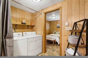A room with a wooden interior, a washing machine, a bed with a checkered bedspread, and a shelf with toiletries. - Rainbow Falls Cabin Sauna Hot Tub Foosball (Sapphire)