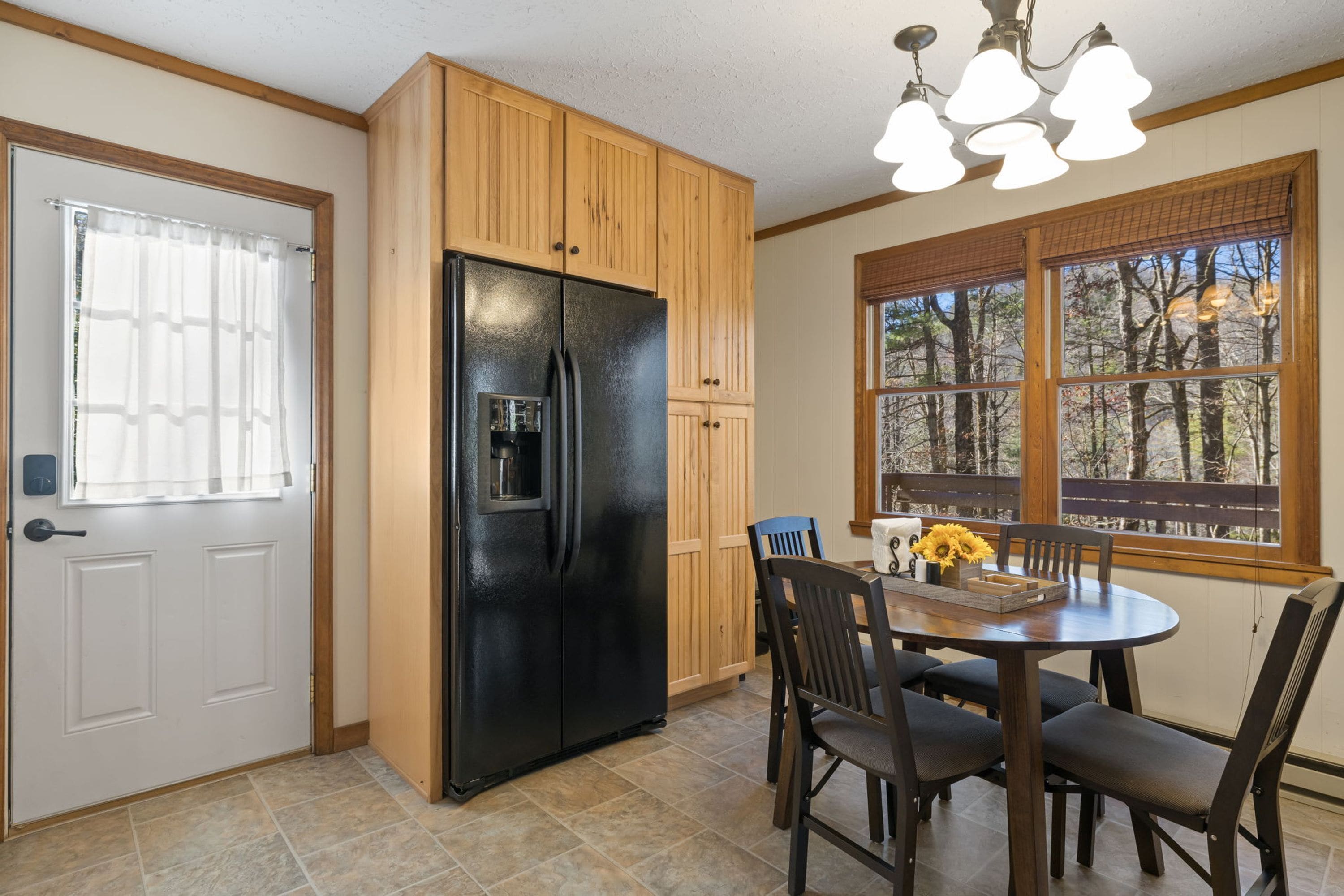A kitchen with a black refrigerator, wooden cabinets, a dining table with chairs, and a window with a view of trees.