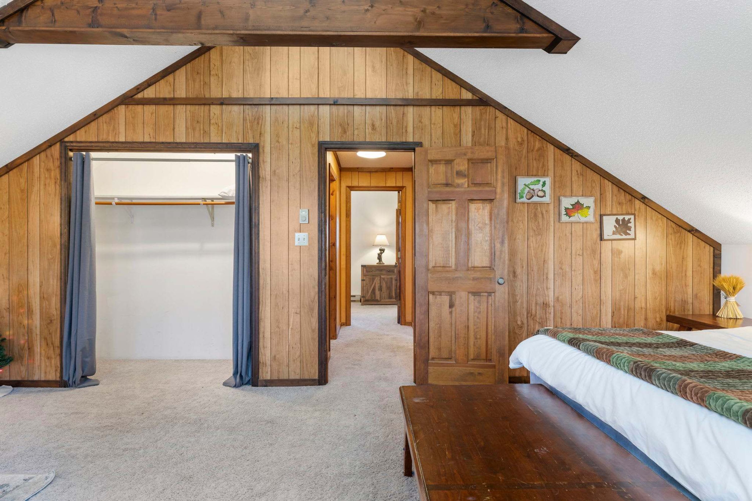 A bedroom with a sloped ceiling, wooden walls, a bed, a bench, and a closet with a curtain.