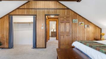 A bedroom with a sloped ceiling, wooden walls, a bed, a bench, and a closet with a curtain.