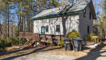 A small, single-story cabin with a wooden deck and a covered area, surrounded by trees.