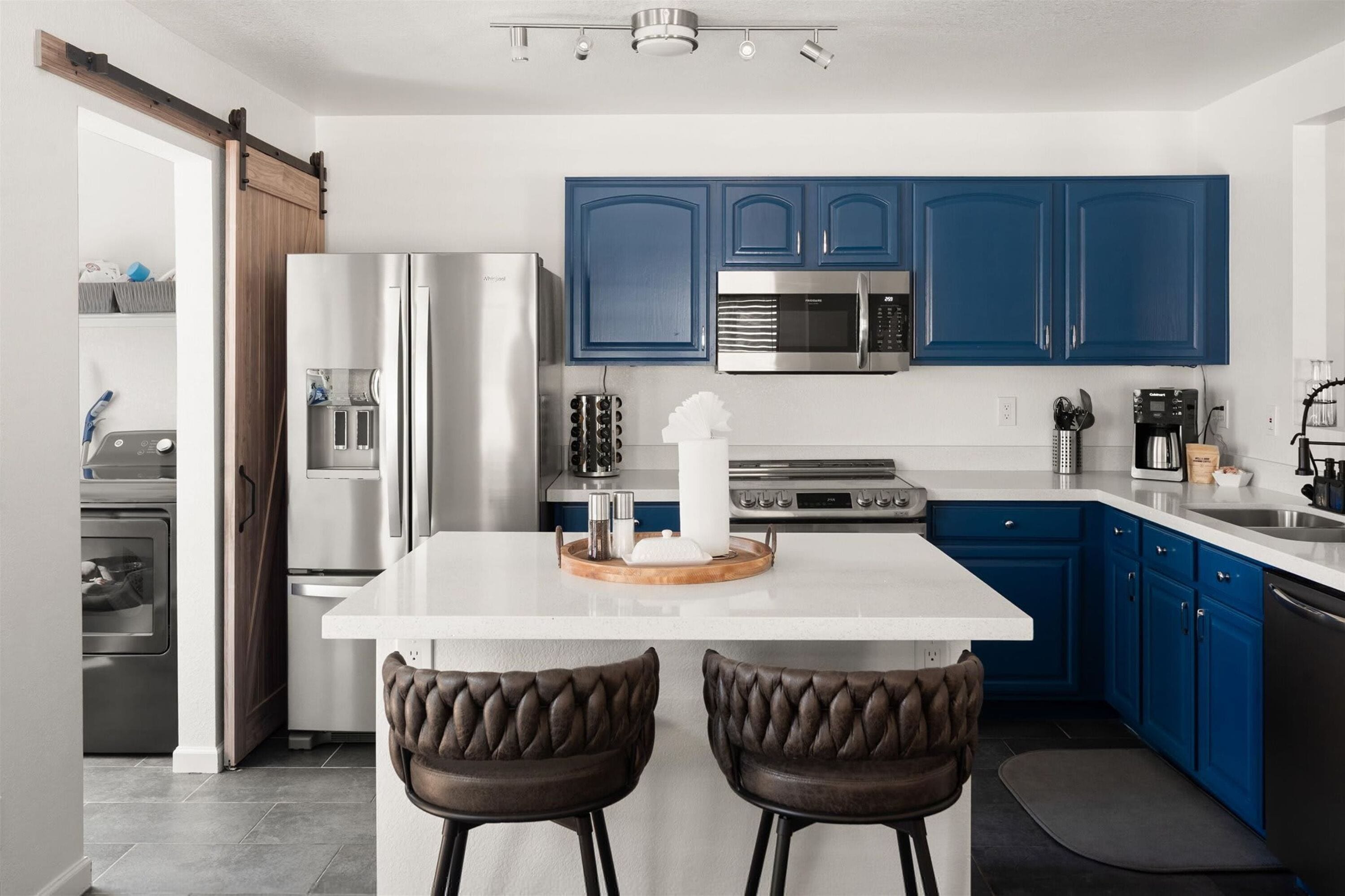 A modern kitchen with blue cabinets, stainless steel appliances, and a central island with two leather stools.