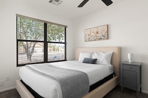 Un dormitorio con cama, mesita de noche, ventana con persianas y un cuadro colgado en la pared.