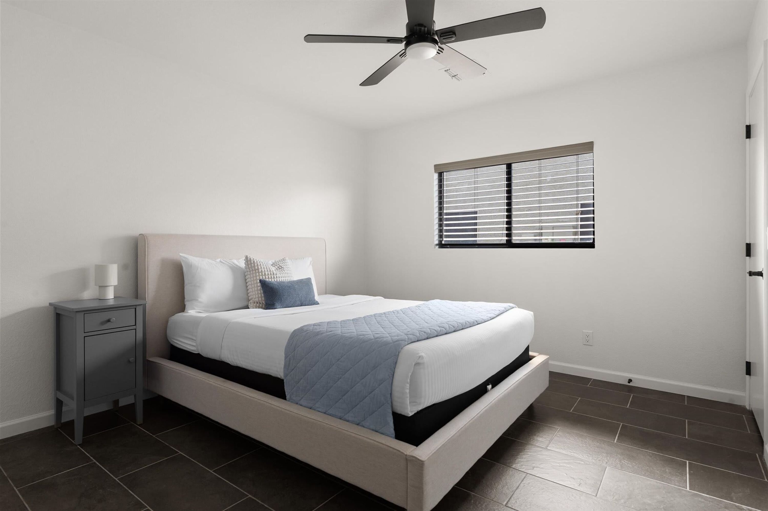 A bedroom with a bed, a nightstand, a ceiling fan, and a window with blinds.