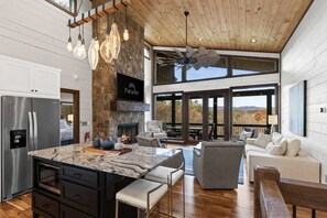 A spacious kitchen with a stone fireplace, a large island, and a modern ceiling fan. - Ellijay Mountain House Hot Tub Views (Ellijay)
