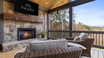 A stone fireplace with a fire, wicker furniture, and a view of trees and mountains.