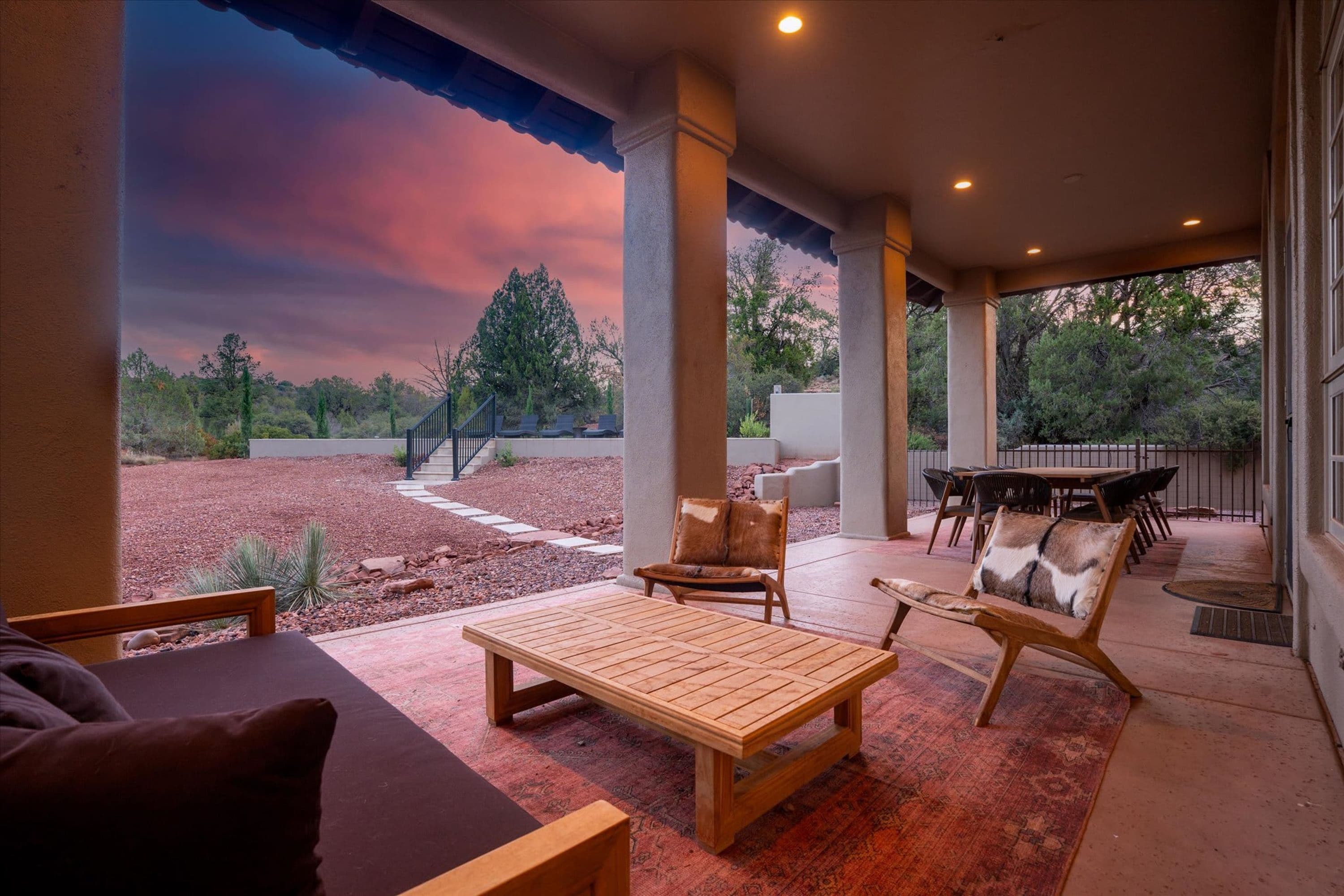 An outdoor patio with a wooden table and chairs, a covered seating area, and a view of a landscaped garden.