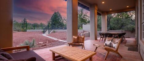 Un patio exterior con una mesa y sillas de madera, una zona de estar cubierta y vistas a un jardín ajardinado.