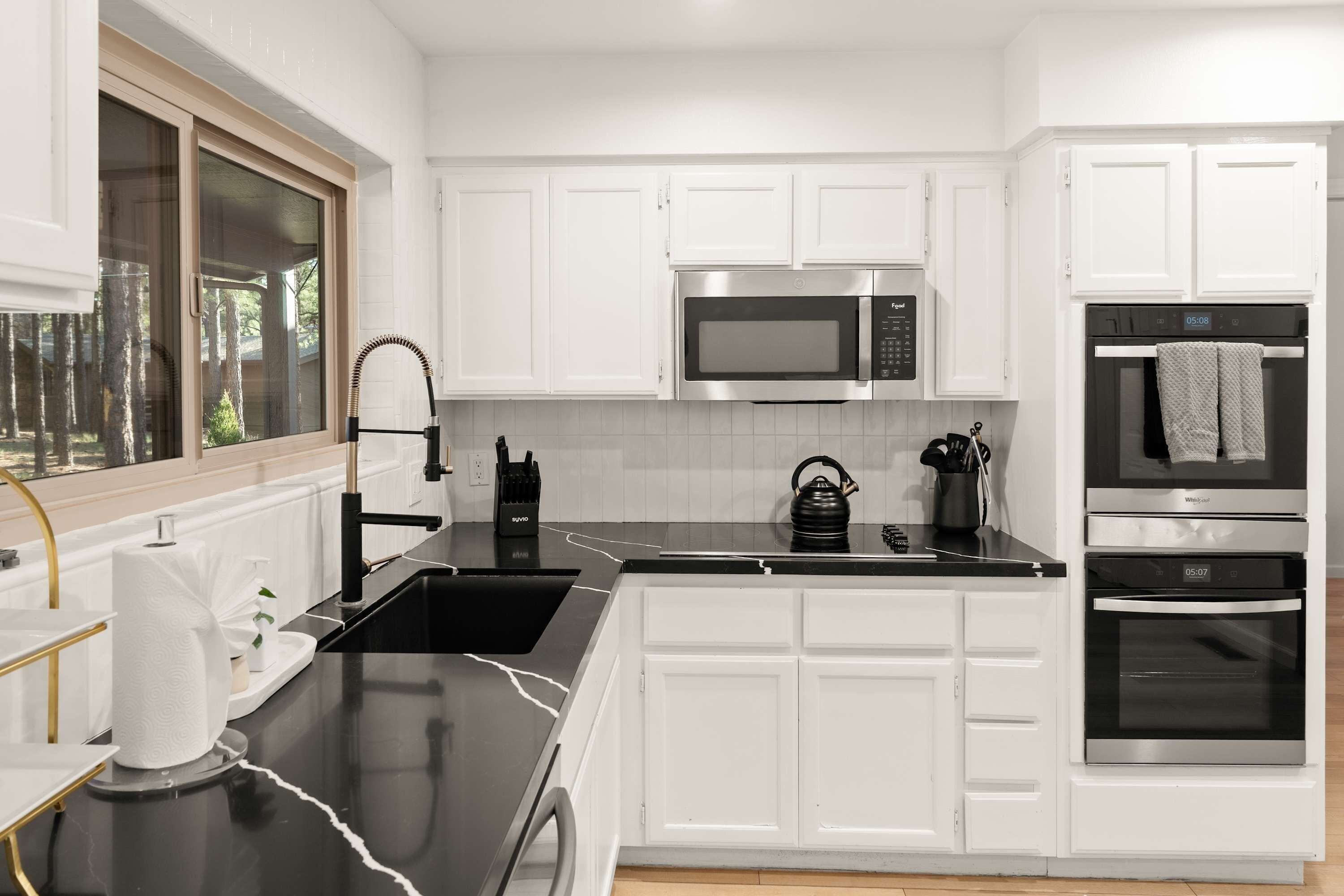 A modern kitchen with white cabinetry, a black countertop, a stainless steel microwave, a kettle, and an oven.
