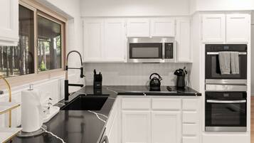 A modern kitchen with white cabinetry, a black countertop, a stainless steel microwave, a kettle, and an oven.