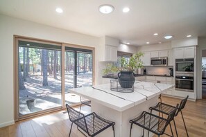 A modern kitchen with white cabinets, a large island with bar stools, and a view of a forest through large sliding doors. - Family-friendly Getaway Hot Tub View EV Chrg (Flagstaff)