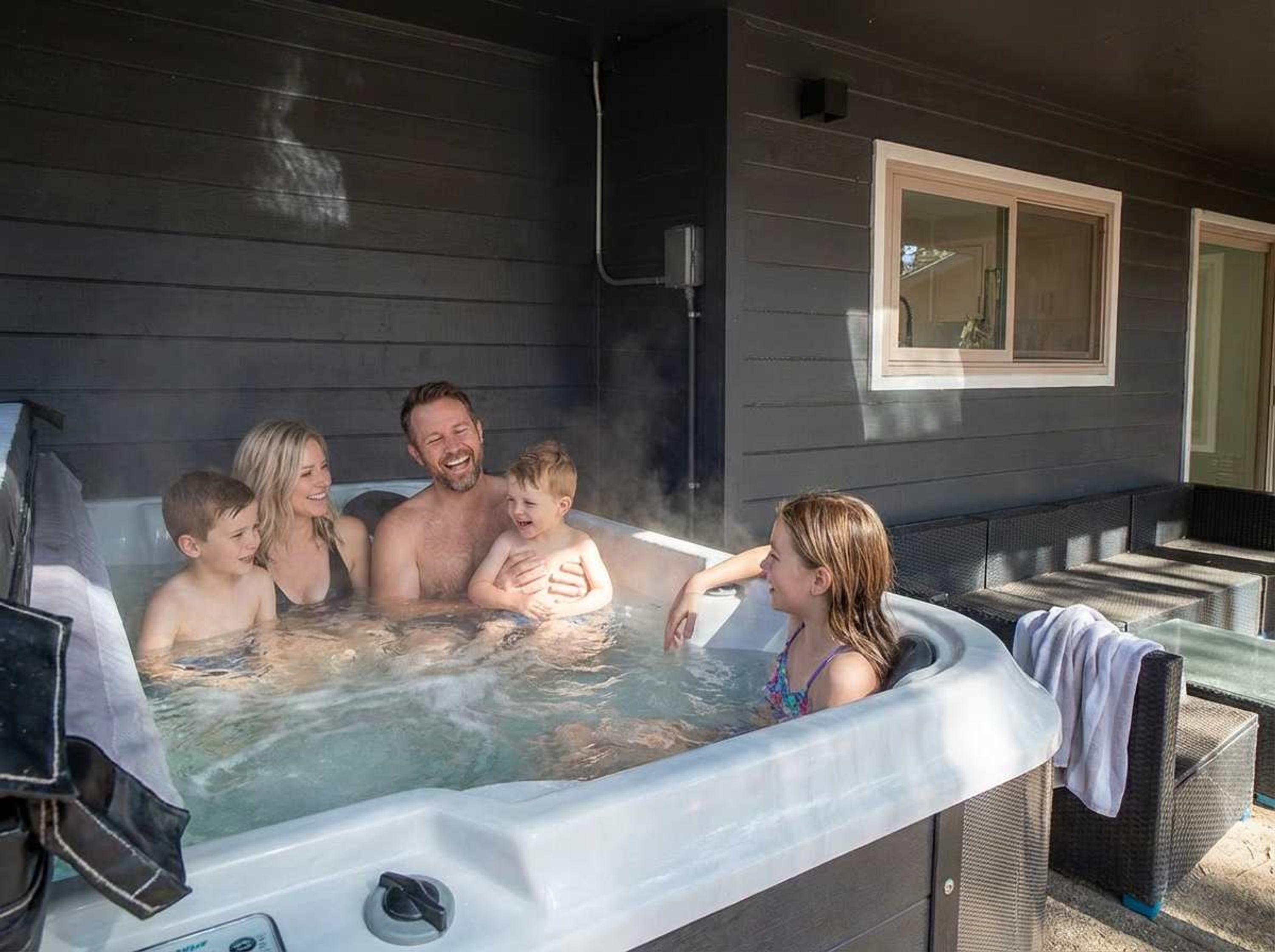 A family of four in a hot tub outside a house with a dark exterior and a window.