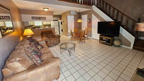 Un salon spacieux comprenant un canapé, un fauteuil et un coin repas avec des chaises. Un téléviseur est posé sur un meuble, et un escalier mène à l’étage supérieur.
