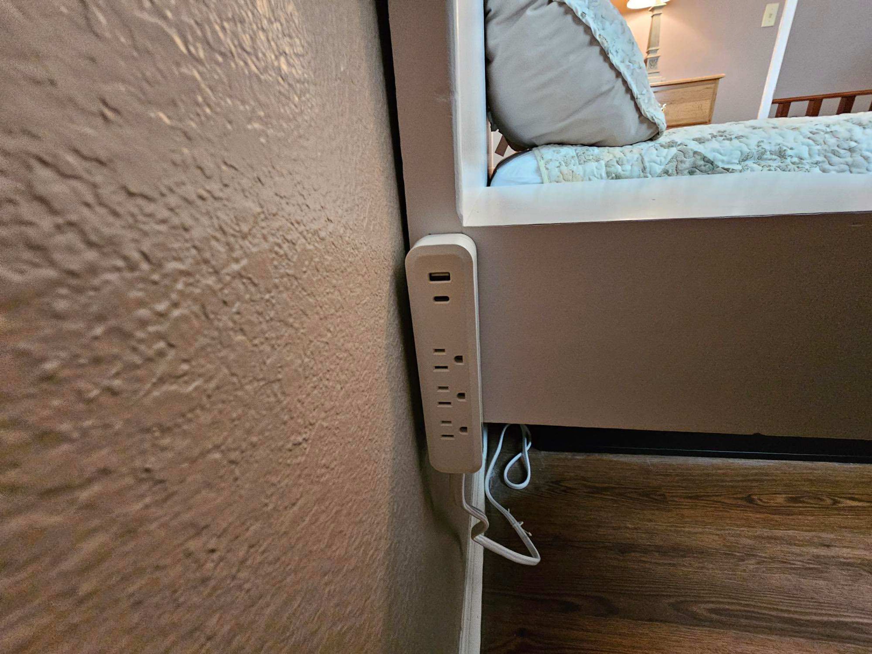 Una cama con cabecera frente a una pared con textura, una mesita de noche con lámpara y un enchufe con cable.