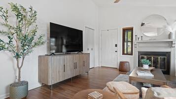 A modern living room with a flat-screen TV on a wooden cabinet, a large potted plant, and a fireplace with a mirror above it.