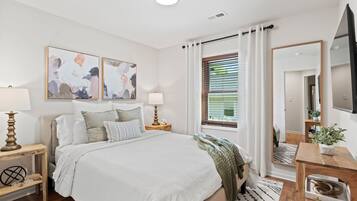 A modern bedroom with a large bed, two bedside tables with lamps, abstract wall art, a window with blinds, a mirror, and a TV on the wall.