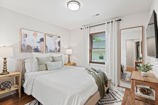 A modern bedroom with a large bed, two bedside tables with lamps, abstract wall art, a window with blinds, a mirror, and a TV on the wall. - 1 5 Miles to Mizzou Spacious Updated Home (Columbia)