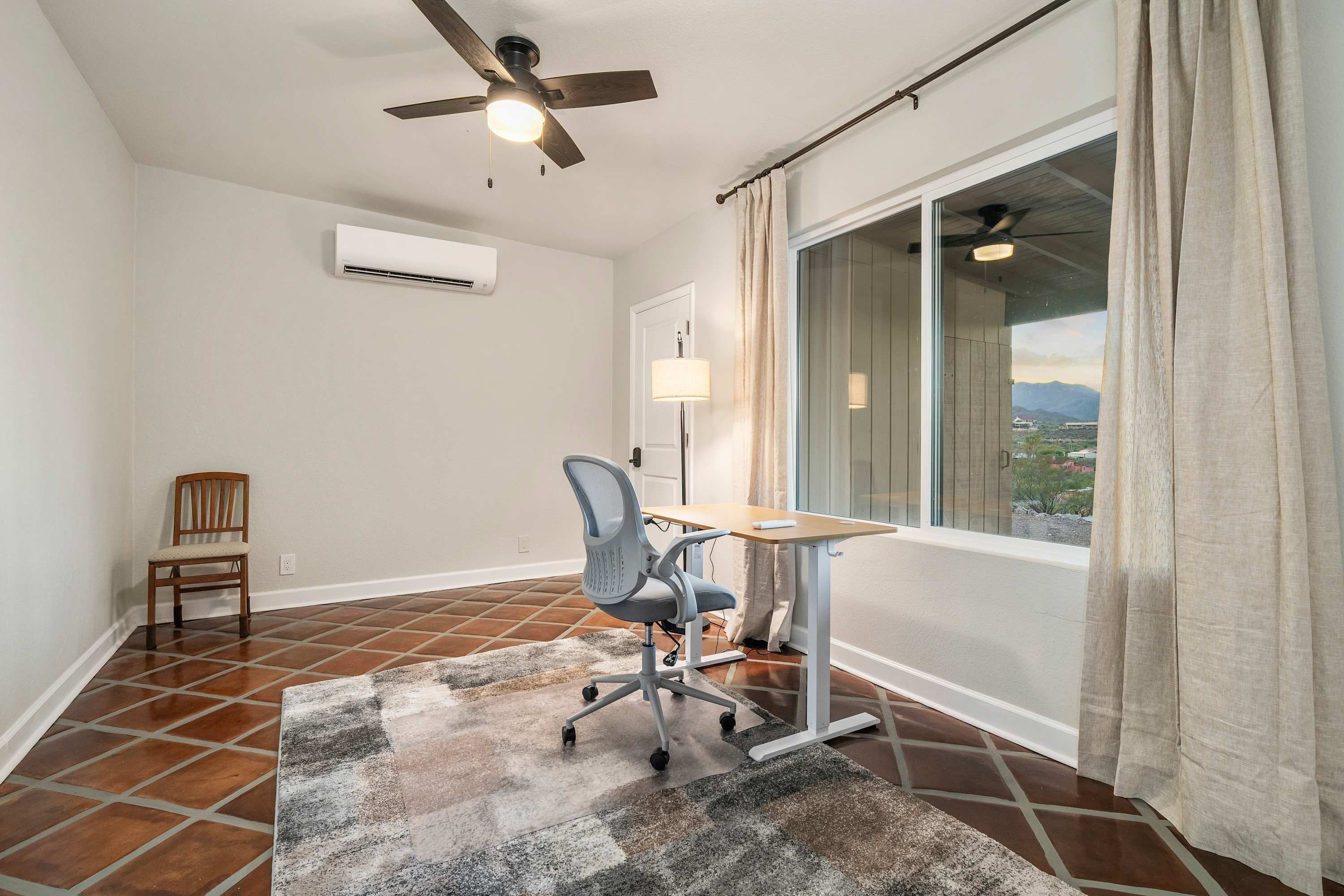 A room with a ceiling fan, an air conditioning unit, a desk with a chair, a window with curtains, and a view of the outdoors.