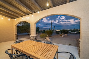 Un patio al aire libre con una mesa y sillas de madera, aberturas en arco y luces colgantes.