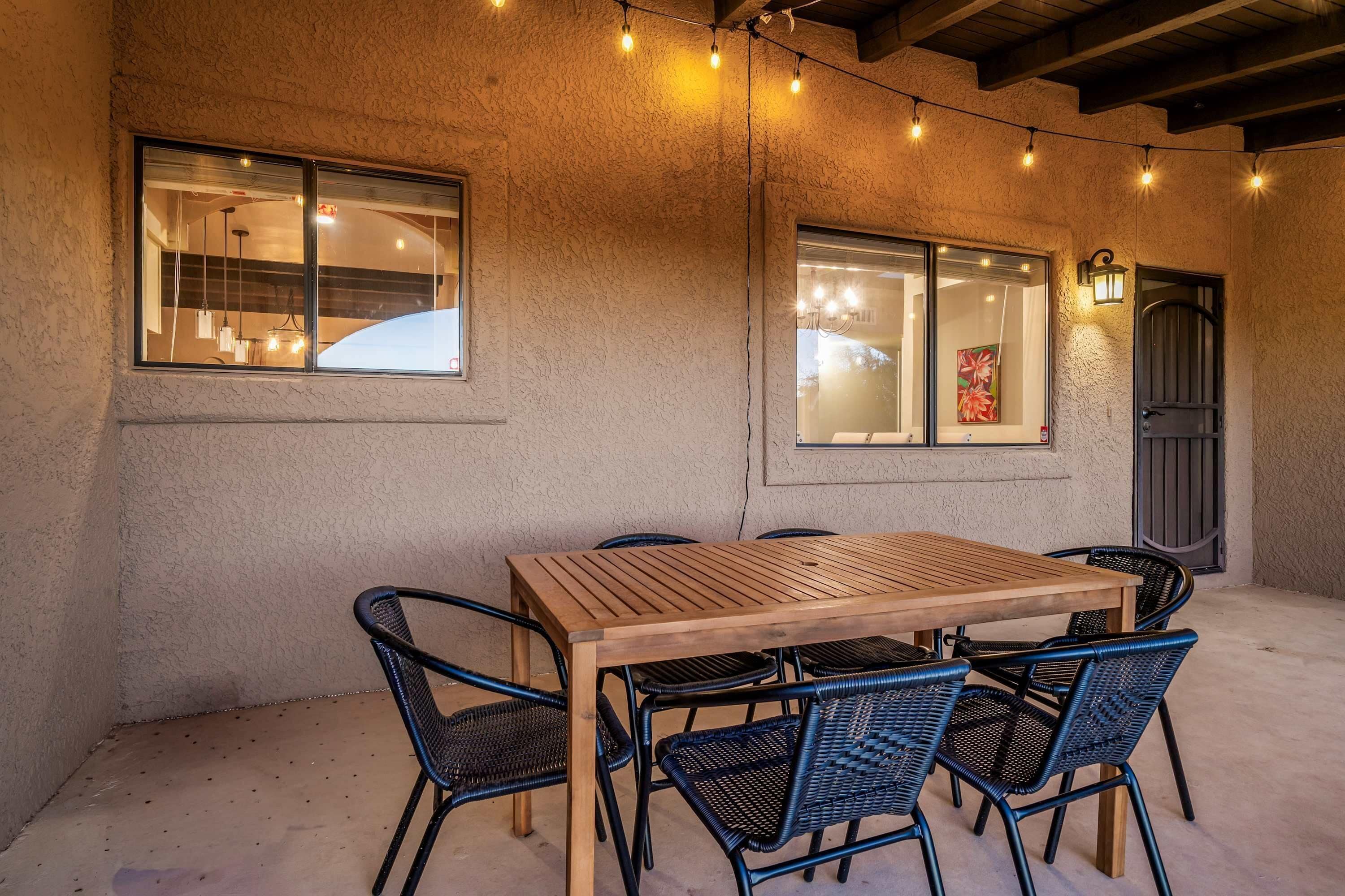 An outdoor dining area with a wooden table and black wicker chairs, string lights, and a textured wall.