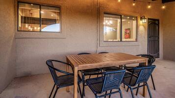 An outdoor dining area with a wooden table and black wicker chairs, string lights, and a textured wall.