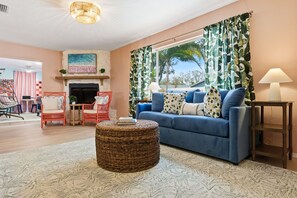 Sala de estar con un sofá azul, chimenea, lámpara colgante y un ventanal con cortinas tropicales.