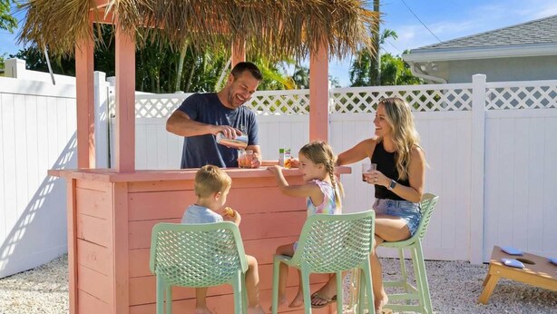 Una família de quatre persones a un petit bar exterior amb teulada de palla, envoltat per una tanca blanca i palmeres.