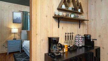 A hotel room with a bed, a desk with a lamp, a coffee station with a coffee maker, and a shelf with mugs.