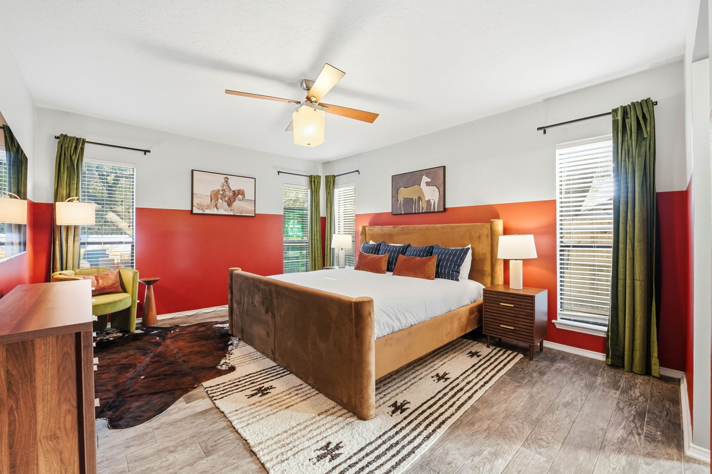 Une chambre à coucher comprenant un lit, une table de chevet, un bureau, une chaise, un ventilateur de plafond et des œuvres d’art accrochées aux murs.